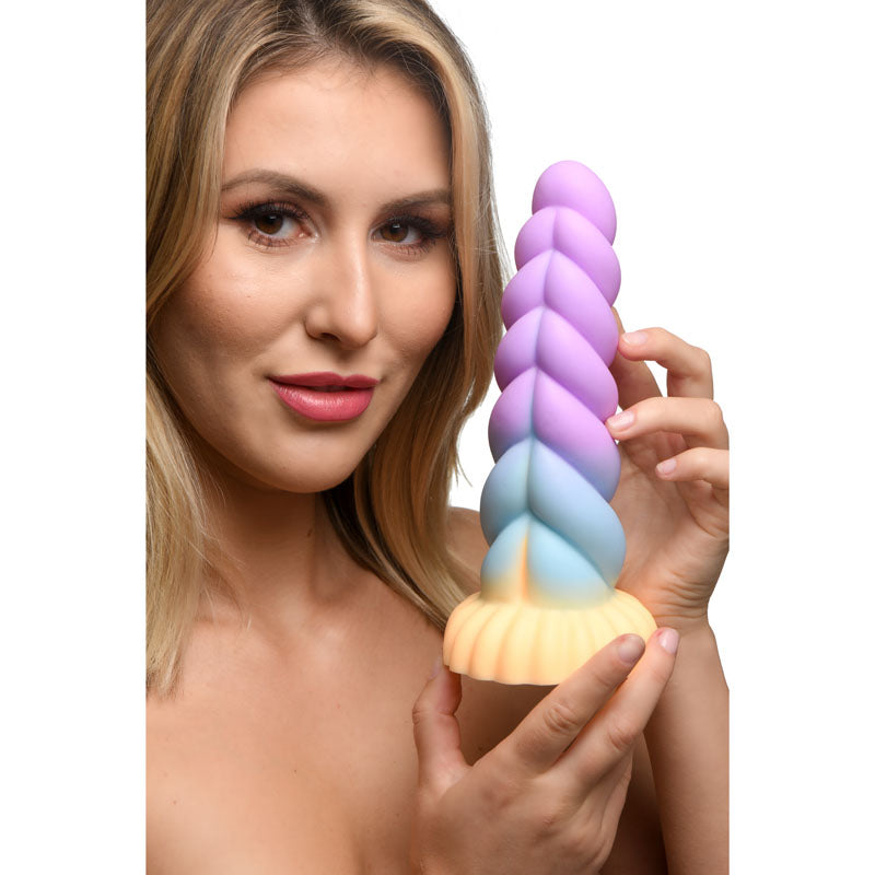 A person with long blonde hair holds the Creature Cocks Mystique Silicone Unicorn Dildo, a 21 cm fantasy dildo with a colorful spiral shape in pastel purple, blue, and yellow. They smile slightly while looking at the camera.
