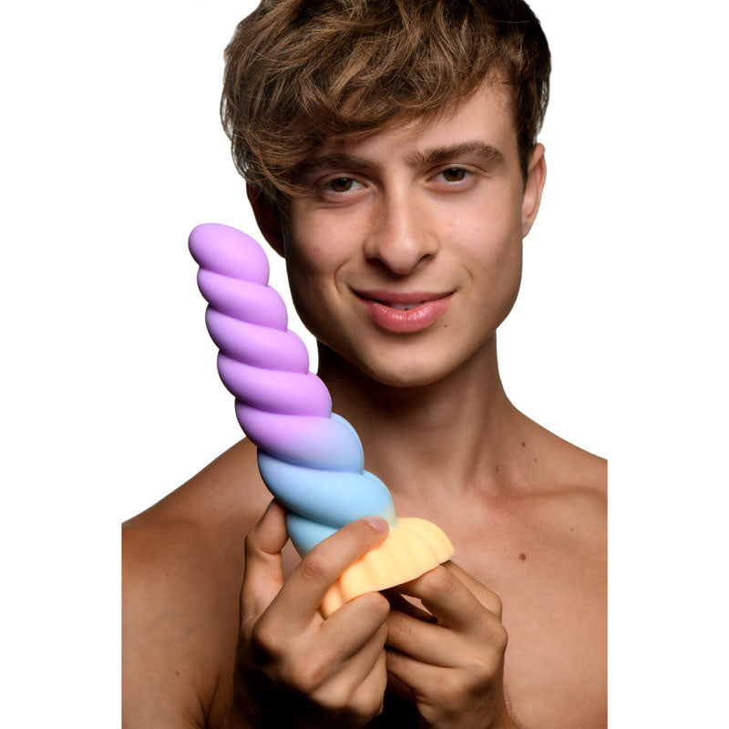 A shirtless person with light brown hair holds a Mystique Silicone Unicorn Dildo from Creature Cocks, its enchanting spiral shape and gradient hues of purple, blue, and yellow. Set against a plain white background, the person smiles softly as theyre mesmerized by this whimsical fantasy dildo.