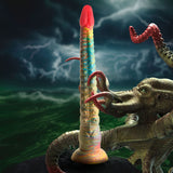 The Creature Cocks Tenta-Dick is a 32 cm multicolored fantasy dildo crafted from phthalate-free silicone, featuring pink to blue and green gradients with ridges and bumps. It stands against a stormy sea backdrop, surrounded by smaller tentacles and illuminated by lightning flashes.