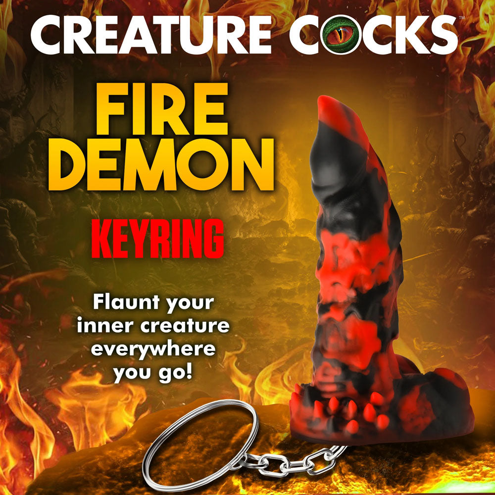 A promotional image for the Creature Cocks Fire Demon Keychain - Novelty Fantasy Dildo Keyring features a fiery red and black demon design against a blazing, hellish backdrop with text: Flaunt your inner creature everywhere you go with Creature Cocks keychains!.