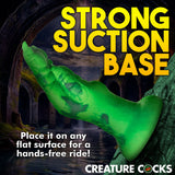 The Creature Cocks Raptor Claw Fisting Silicone Dildo in green, featuring a strong suction base angled upward like a mighty claw, is set against a dark tunnel backdrop. Bold yellow text states STRONG SUCTION BASE, with white text advising, Place it on any flat surface for hands-free fantasy roleplay!.