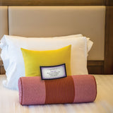 A hotel bed with crisp white sheets is brightened by a yellow pillow and a pink bolster with red stripes, sparking tantalizing fantasies. Beneath the elegant wooden headboard, an Adventurous Sex Coupons set rests on the bolster alongside a framed card about cleanliness.