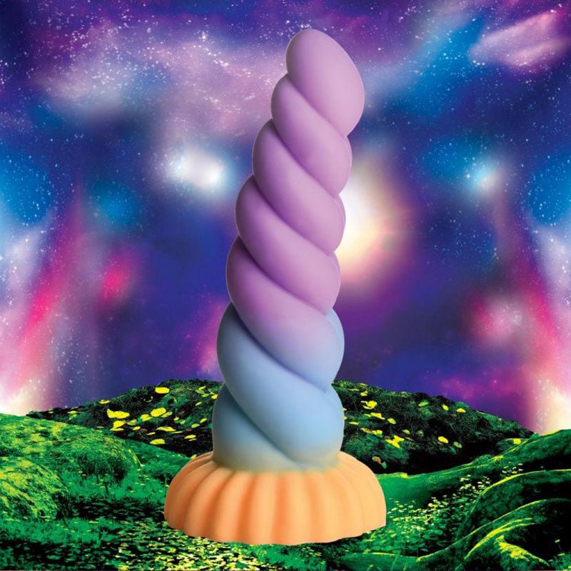 The Creature Cocks Mystique 21 cm Silicone Unicorn Dildo features a vibrant purple and blue spiral design atop an orange, scalloped suction cup base, set against a starry sky with glowing clouds and a lush landscape accented by yellow highlights.